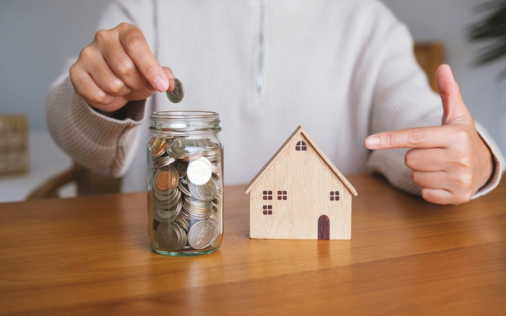 oman putting coins into a glass jar and pointing finger at a wooden house model for saving money